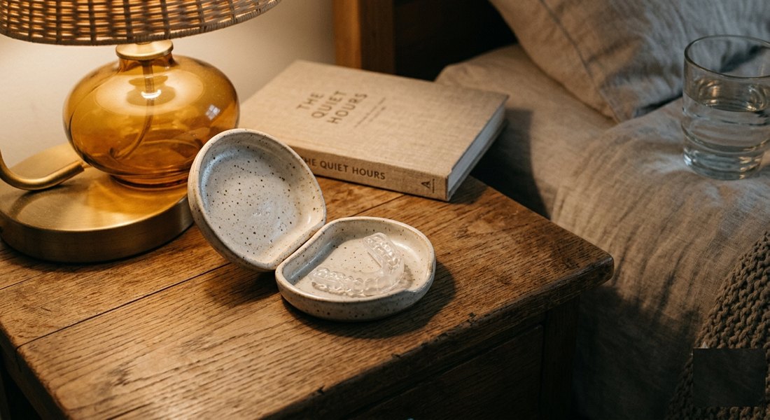 A clear dental guard in an open ceramic case on a lamp-lit bedside table